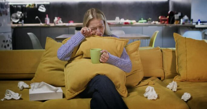 Woman cries embracing pillow with cup of tea in hands. Lady watches soap opera reacting emotionally on plot of movie wiping tears with tissues
