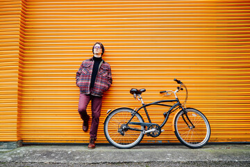 Young stylish hipster man standing with a cruiser city Bicycle near orange Wall. Student Resting...