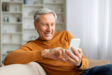Happy elderly man resting on couch at home, using smartphone