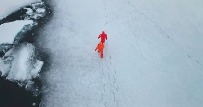 Two man in immersion suits walk Antarctica ocean ice. People explore South Pole. Group of scientists in orange wetsuits research climate and arctic wild nature. Aerial top trekking drone shot