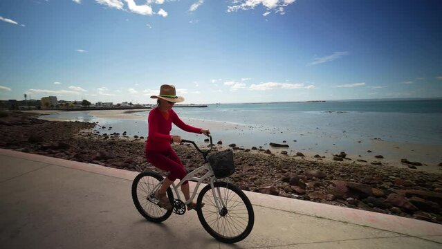 Profile view of mature woman riding a bike on the La Paz malecon cyclovia bicycle trail along the waterfront with palm trees.