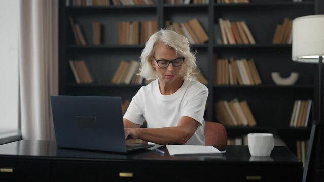 Senior woman freelancer works on laptop. Mature gray haired female in glasses working at home office writing notes typing on computer. Confidence expert doing her job. Freelance online distant work.