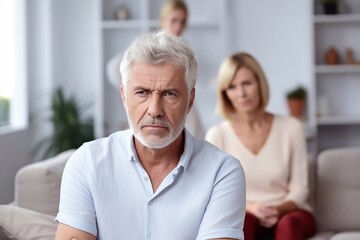 Fototapeta na wymiar Senior caucasian spouses misunderstanding each other, experiencing crisis in relationship, offended wife looks disappointedly at camera, husband sits in defocus on background. Quarrel, marital crisis
