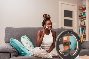Joyful African Woman sitting on the couch Enjoying Skincare Routine During Vlogging Session in...