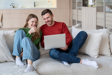 Obraz premium Glad joyful married couple laughing happily, looking into laptop computer screen. Cheerful wife and husband staying at home during lockdown, spending time together, watching comedies, funny videos