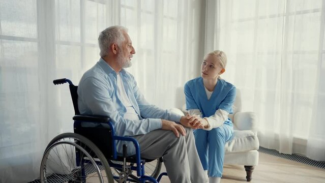 Nurse woman cares old man in wheelchair. Female social welfare worker physical therapist looks after senior patient providing medical care at home. Living with legs paralysis, special needs people.