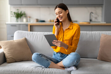 Smiling young woman multitasking with phone and tablet on sofa