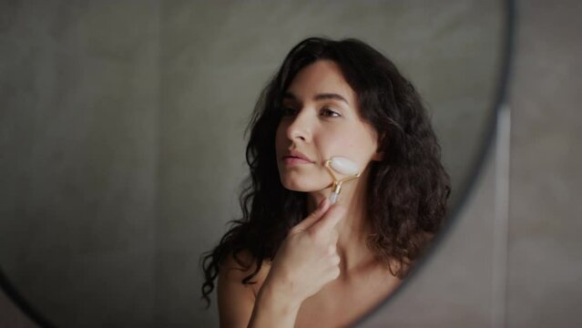 Close-up shot of mirror reflection of beautiful smiling Caucasian woman with long dark curly hair and bare chest standing in bathroom and doing face massage with gua sha roller