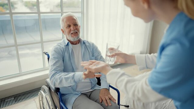 Nurse gives old wheelchair man pills. Physical therapist woman provides help support to senior paralyzed male gives water medications. Palliative care of neurodegenerative pathologies concept.