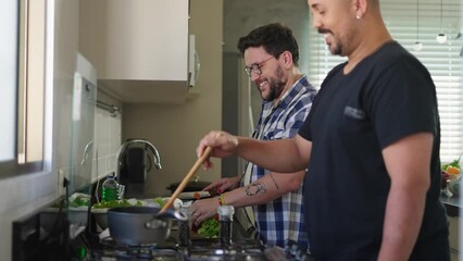 A cheerful male gay couple preparing lunch together in the kitchen. Happy attractive young adult men joyfully spending time together cooking delicious dishes side by side.
