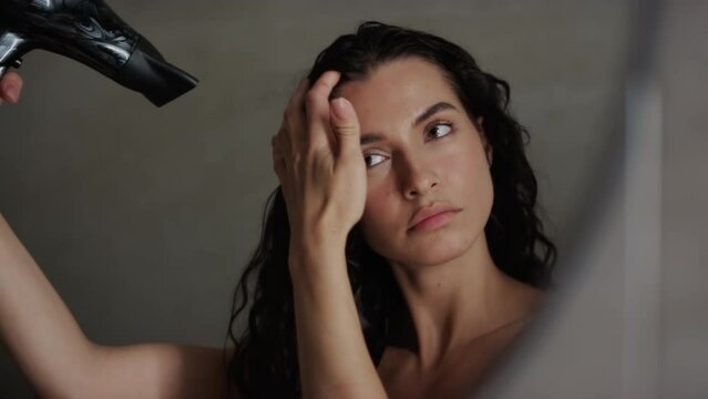 Close-up shot of mirror reflection of beautiful Caucasian woman with long curly hair, bare shoulders standing in bathroom and using hair dryer after washing in shower