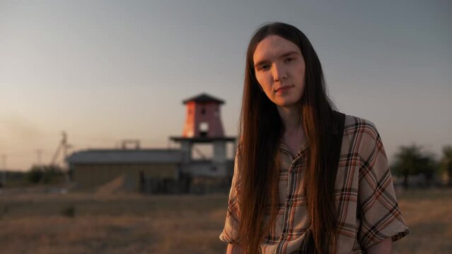 Lonely sad lgbt guy with long straight hair standing on a farm in the setting sun looking sadly at the camera 