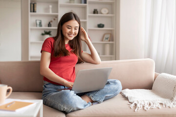 Happy young asian woman comfortably sitting on couch and working on laptop