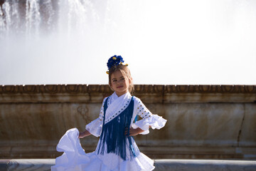 A pretty little girl dancing flamenco dressed in a white dress with ruffles and blue fringes in a...
