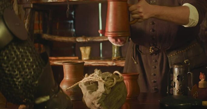 Attendant pours drink to knight mug in tavern closeup. Careful innkeeper gives jug of wine to warrior in armour at medieval inn. Adventurer visits pub