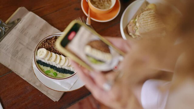 Female hands taking photos of smoothie bowl, menu on table by smartphone. Tasty sandwich, coffee on table. Woman photographs healthy food in hipster cafe on Bali. Blogger takes photo of the breakfast.