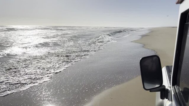 Sandwich Harbour, Namibia - August 22, 2022: A rugged Land Rover Defender driving along the beach, just a few meters from the water. Shot recorded from the left rear window.