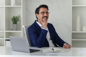 Naklejka premium Thoughtful businessman in a blue suit sits at his desk with a laptop and notebook, hand on chin