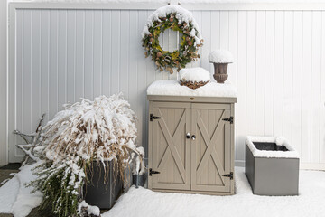 Snow covers the patio decoration like a little cabinet, a green wreath, an amphora and flowerpots