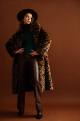 Fashionable confident woman wearing hat, trendy long faux fur leopard print coat, green turtleneck, leather pants, pointed toe boots, posing on brown background. Full-length studio fashion portrait