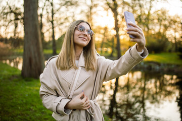 Stylish beautiful young girl in sunglasses and a beige trench coat takes a selfie against the backdrop of trees and a river lake.