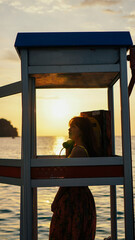 Portrait of a charming, bright, and happy girl posing in a vintage phone booth on a sea background....