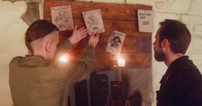 Rogues choose beast to hunt on bulletin board in tavern. Friends tramps discuss profitable task in medieval pub. Adventurers seek good job in bar