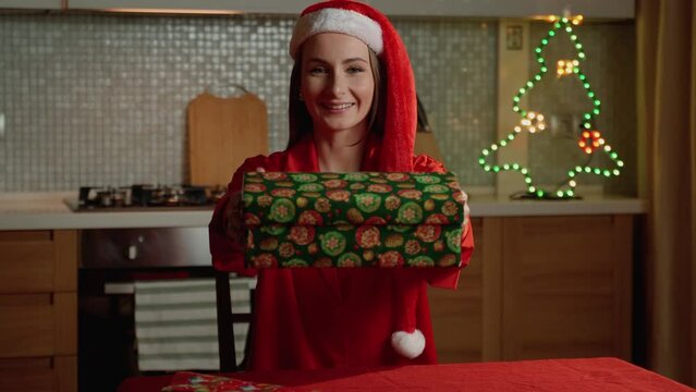 A joyful woman in a Santa hat proudly holds out a wrapped present, with twinkling Christmas lights in the background enhancing the holiday atmosphere.