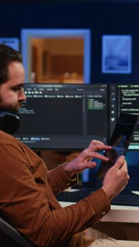 Software developer programming in home office, crosschecking lines of code between tablet and PC screen. IT admin holding portable device, using it as reference while coding on computer, camera B
