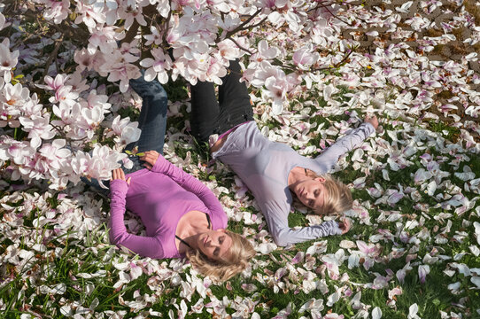 Identical twin sisters lying on grass under a magnolia tree surrounded by magnolia petals