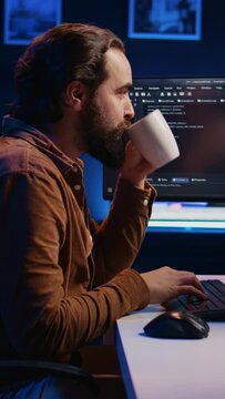 Focused programmer drinking coffee in apartment while solving database errors on computer. Remotely working IT admin concentrated on finalizing coding project in home office, camera B