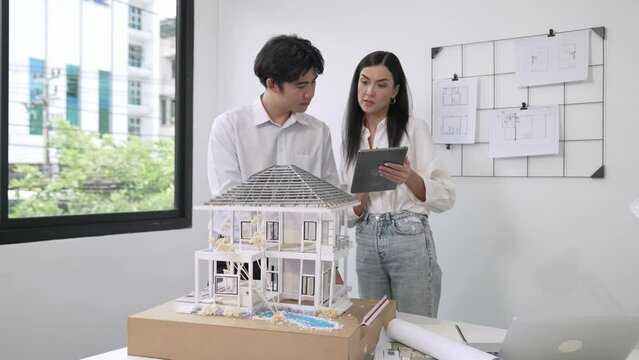 Beautiful young caucasian project manager brief asian engineer by using tablet while engineer measuring house model on meeting table with blueprint, architectural equipment scatter around. Immaculate