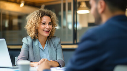 A smiling woman with glasses is engaged in a conversation with a male colleague in a modern office setting, creating an atmosphere of friendly professional interaction.