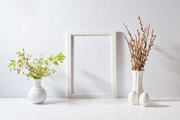 Mockup with a white frame and spring flowers in a vase, easter eggs on a light background. Empty...