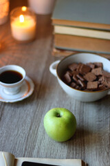 Vintage books, e-reader, pen, reading glasses, phone, cup of tea or coffee, bowl of cookies, green apple and lit candles on the table. Hygge at home, studying atmosphere. Selective focus.