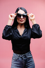 Young caucasian girl looking at camera through sun glasses on pink background. Dark-haired beauty wears black shirt. People sincere emotions lifestyle concept.