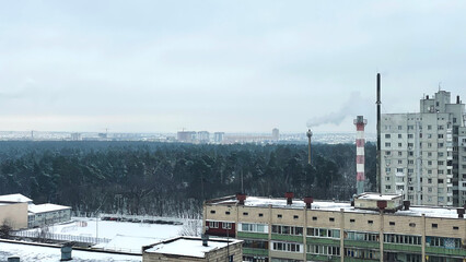 A toxic chimney of an enterprise against the background of the sky and the city releases smoke. The factory pollutes the environment. Industrial cityscape. Winter day. Poisoned air.