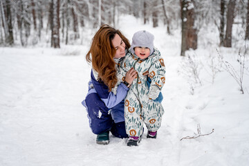 Fototapeta na wymiar A young mother in a warm suit and a little 2-year-old child laugh and have fun in a snowy winter forest on a walk