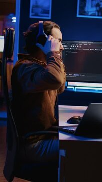 Software programmer taking break from coding, putting headphones on and stretching on desk chair during remote job shift. IT professional pausing tasks, listening music, loosening up, camera B