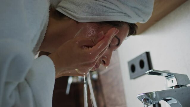 Close-up side shot of young woman with wet hair wrapped in towel, closed eyes, bending over bathroom sink and washing off face mask with water during skincare routine