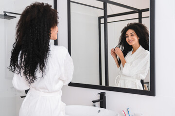 Young african woman female in white bathroom after taking shower bath taking care of her curly hair, applying oil, foam to comb it, preparing for date meeting in the morning