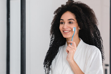 Young african pretty woman in white bathrobe brushing her teeth while looking at the mirror. Daily morning routine, skin care and bathing procedures