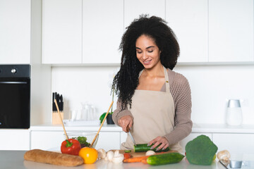 African young woman wearing apron, cooking salad, soup, vegetable vegan vegetarian dishes in the home kitchen, cutting cucumber. Healthy eating habits, dieting concept