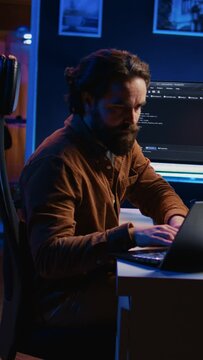 Computer scientist putting headphones on while developing code on PC, making time pass faster during remote job shift. IT admin doing multitasking in home office while listening music, camera B