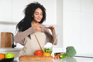 Happy smiley young african woman taking photo vlogging blogging while cooking, recording the...