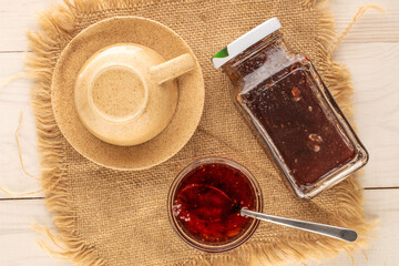 Strawberry jam in glass saucer with metal spoon and glass jar on jute napkin, macro, top view.