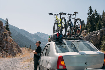 A man stands next to a car with bicycles on the roof on a mountain road.