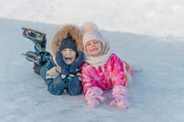 The older brother and younger sister are lying on the ice, with skates on their feet, and laughing. Winter fun.