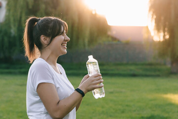 Beautiful girl smiles against the background of the sunset and holds bottle of water in her hand....