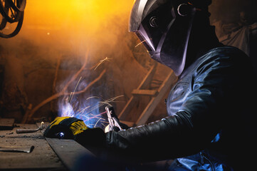 A male welder in a leather jacket carries out welding work in a home shed against the background of a brick wall. Handicraft production of metal structures and products